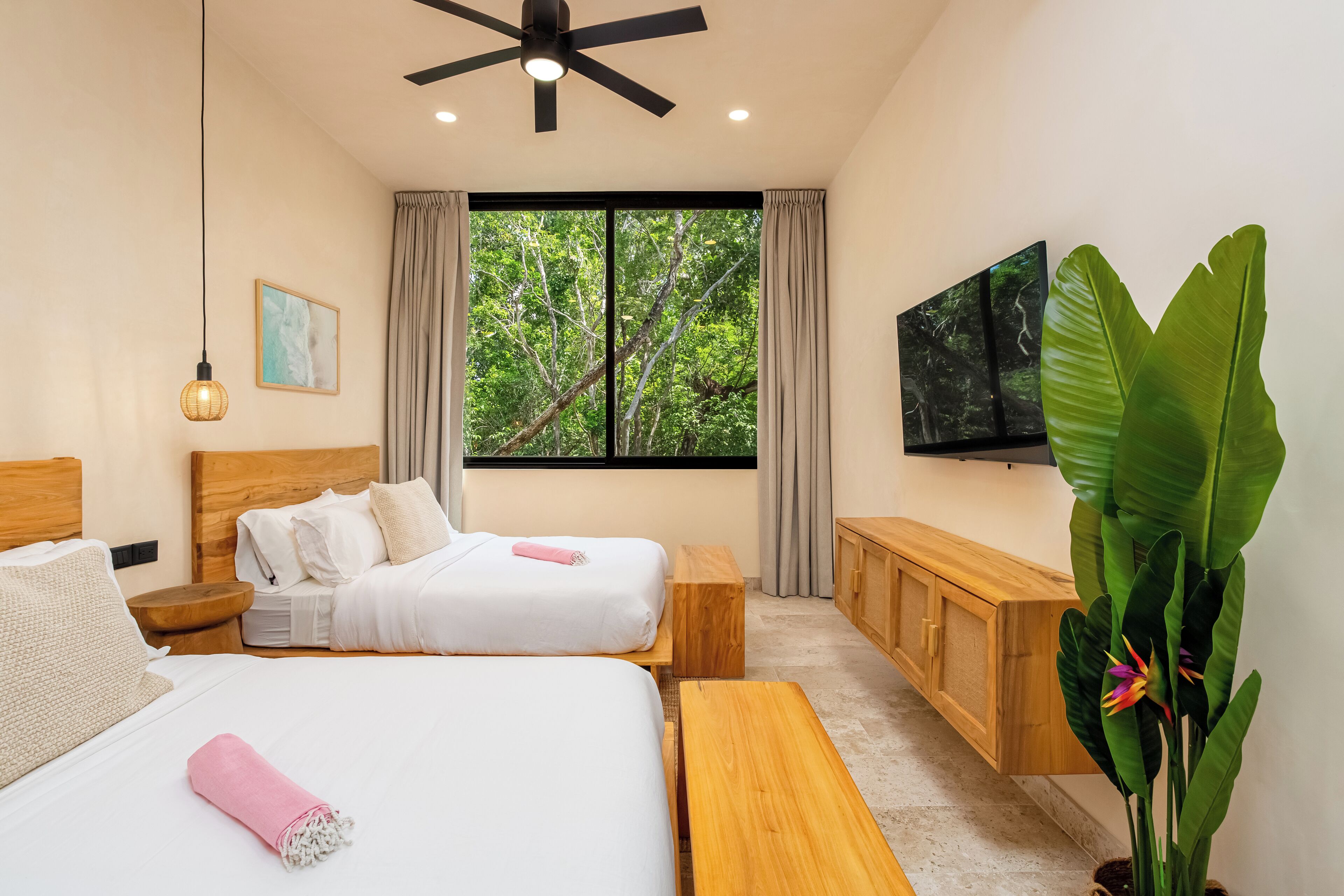 Quarto de hotel com duas camas, um ventilador de teto, uma janela ampla com vista para árvores, um banco de madeira, uma televisão e uma planta grande.