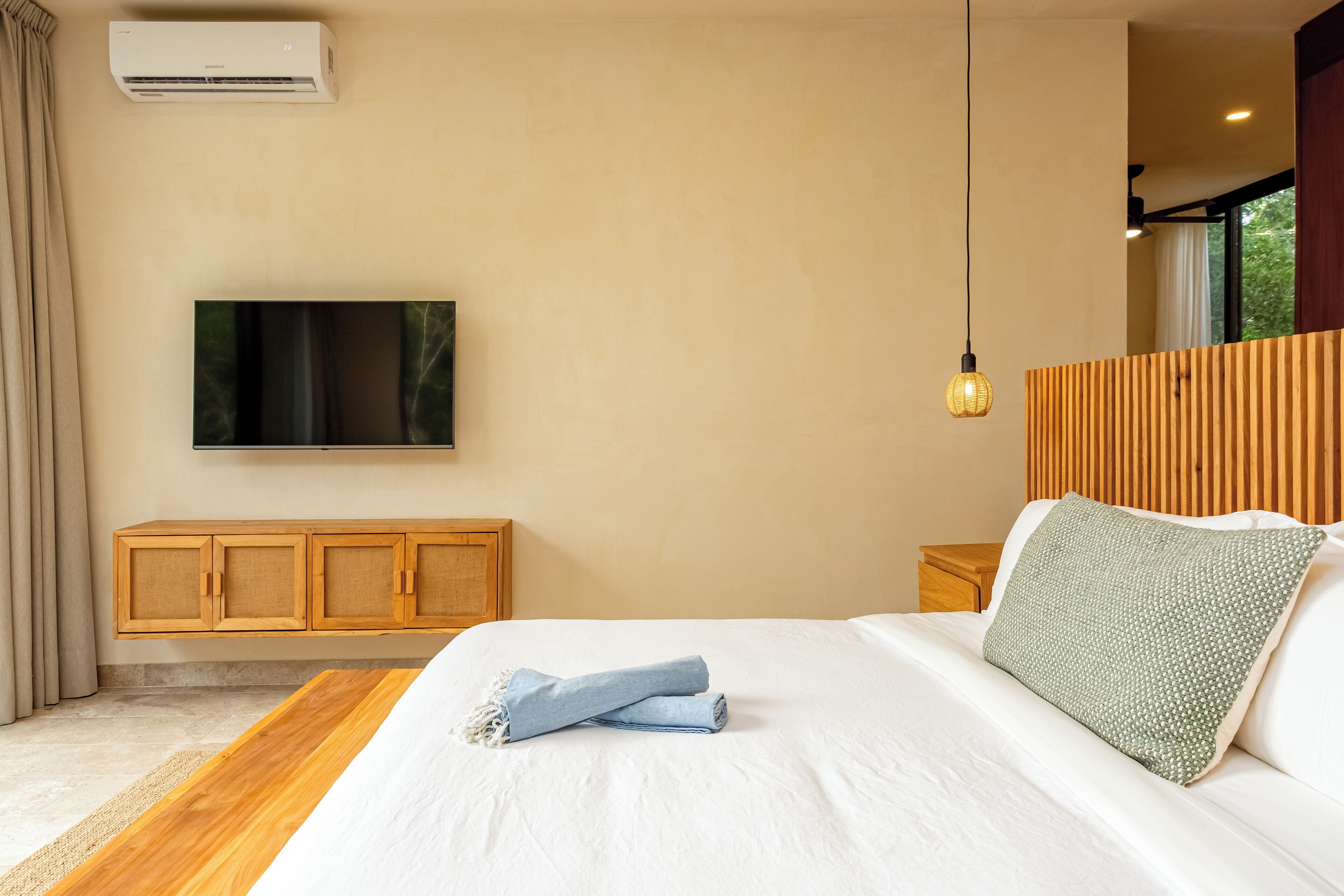A bedroom with a bed, a television mounted on the wall, a wooden headboard, a bedside table, and an air conditioning unit.