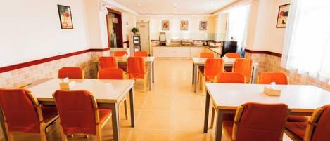 A dining area with tables and chairs, a kitchen in the background, and a tiled floor.