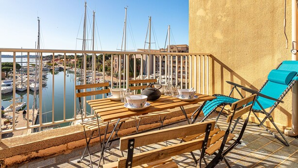 Un balcon agrémenté de meubles en bois, une vue sur une marina avec des bateaux, et une plage de sable.