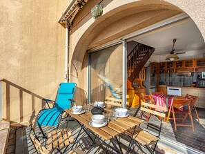 A sunlit patio with a wooden table set for dining, a blue lounge chair, and a view of the kitchen and living area through an open door.