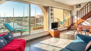 Un vaste espace de vie avec une grande fenêtre donnant sur un port de plaisance, un coffre en bois et un coin salon confortable.