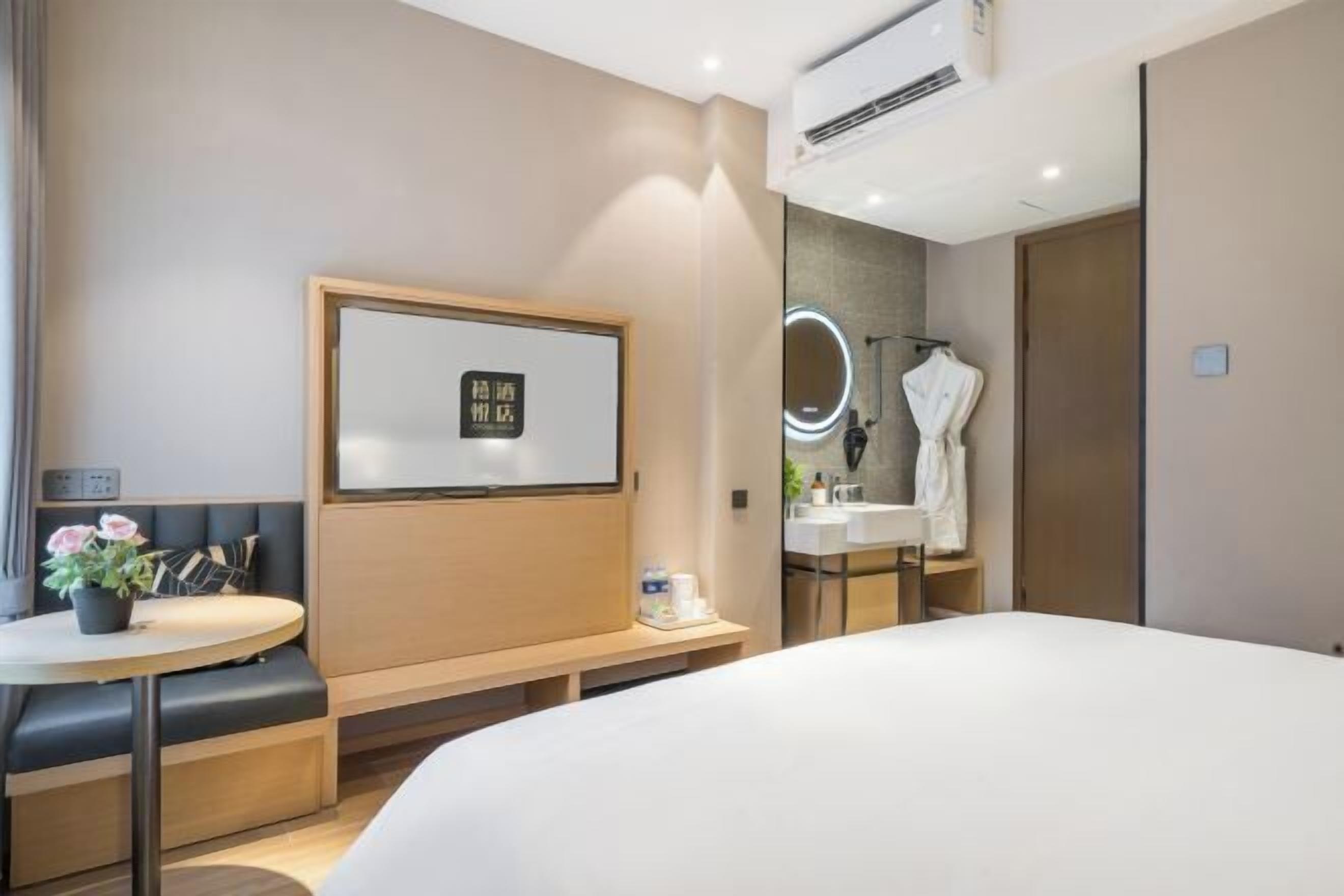 A modern hotel room with a bed, a round table with a potted plant, a bench, a mirror, and a bathroom with a sink and towel rack.