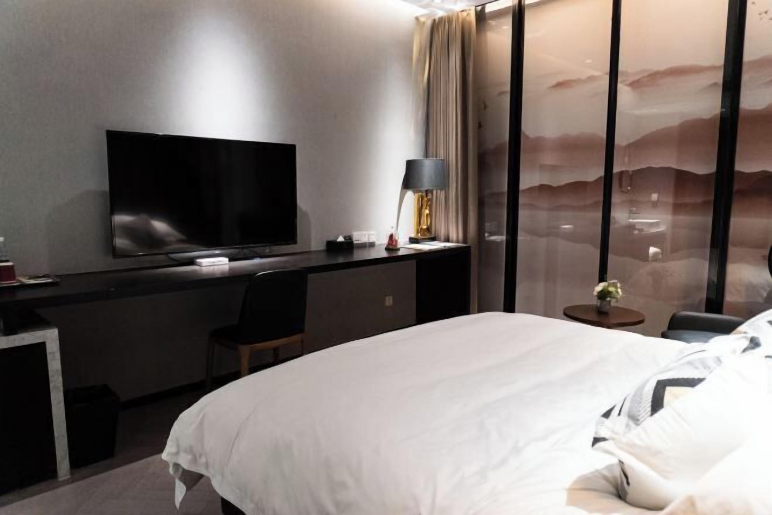 A hotel room with a bed, a desk with a flat-screen TV, a lamp, and a view of a mountainous landscape through a glass partition.