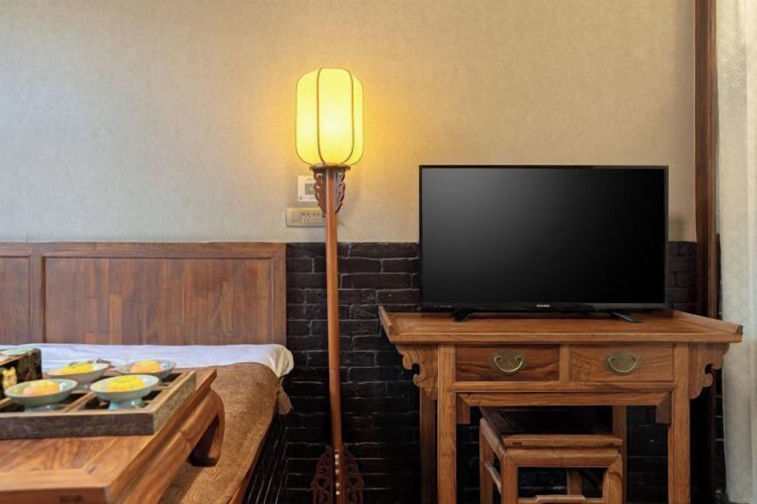 A room with a bed, a television on a wooden stand, a table with plates, and a floor lamp.