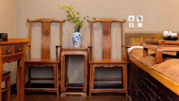 Una habitación con una cama, sillas de madera, una mesita con un florero y un panel de interruptores en la pared.