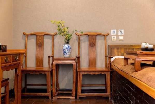 Une pièce comprenant un lit, des chaises en bois, une petite table avec un vase et un tableau de distribution fixé au mur.