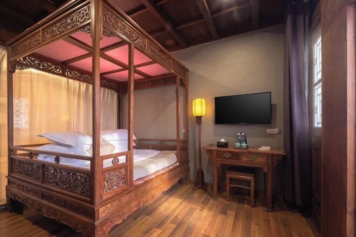 A four-poster bed with intricate carvings, a wooden desk with a lamp, and a flat-screen TV mounted on the wall.