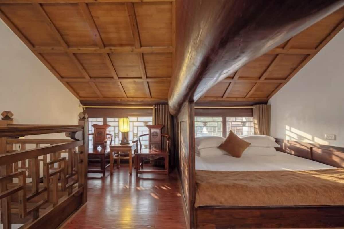 A room with a wooden ceiling, a bed with a brown blanket, a desk with chairs, and a window.