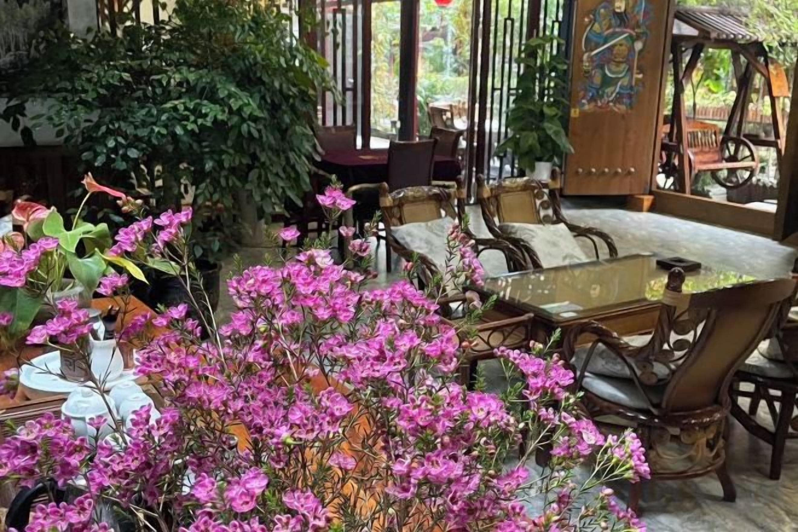 Een kamer met een glazen tafel, houten stoelen en een grote potplant met roze bloemen.