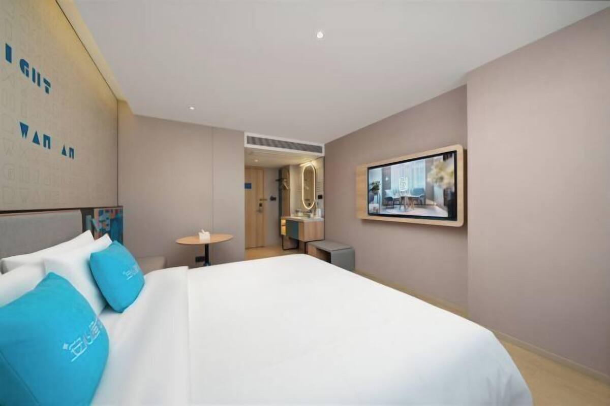 A hotel room with a bed, a desk, a chair, a small table, a mirror, and a framed picture on the wall.