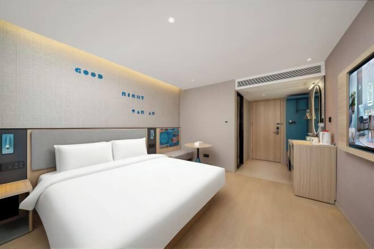 A modern hotel room with a large bed, a bedside table, a small desk, and a mirror.