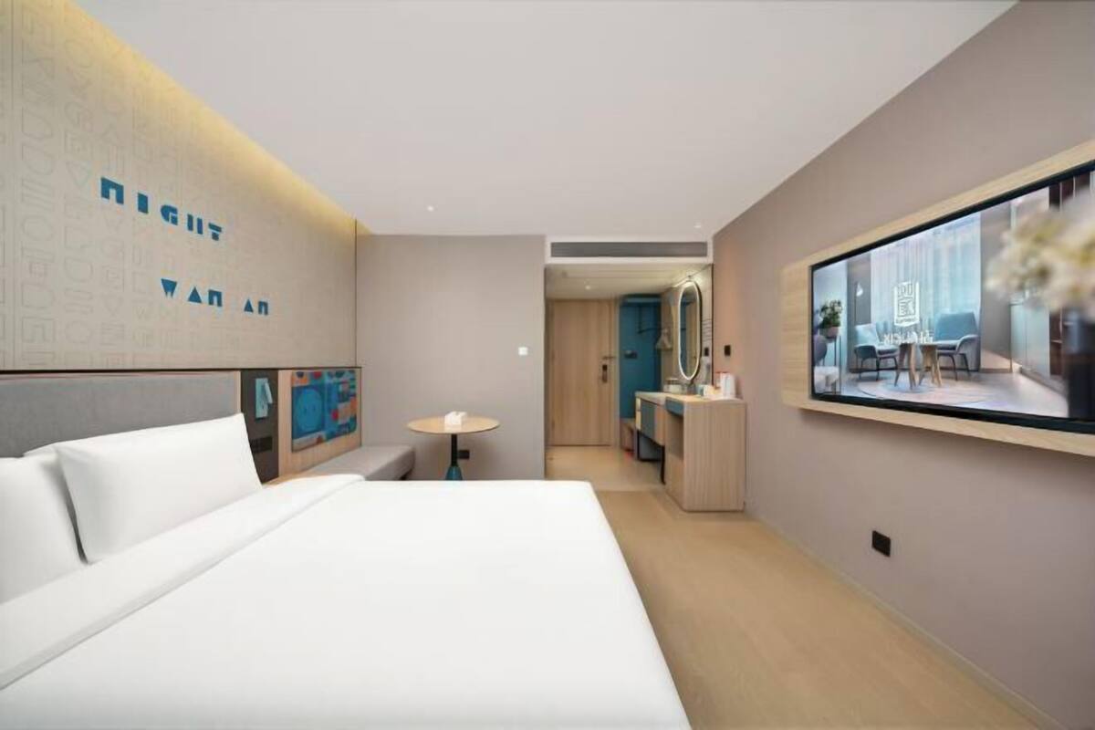 A modern hotel room with a large bed, a desk with a mirror, and a view of the city through a large window.