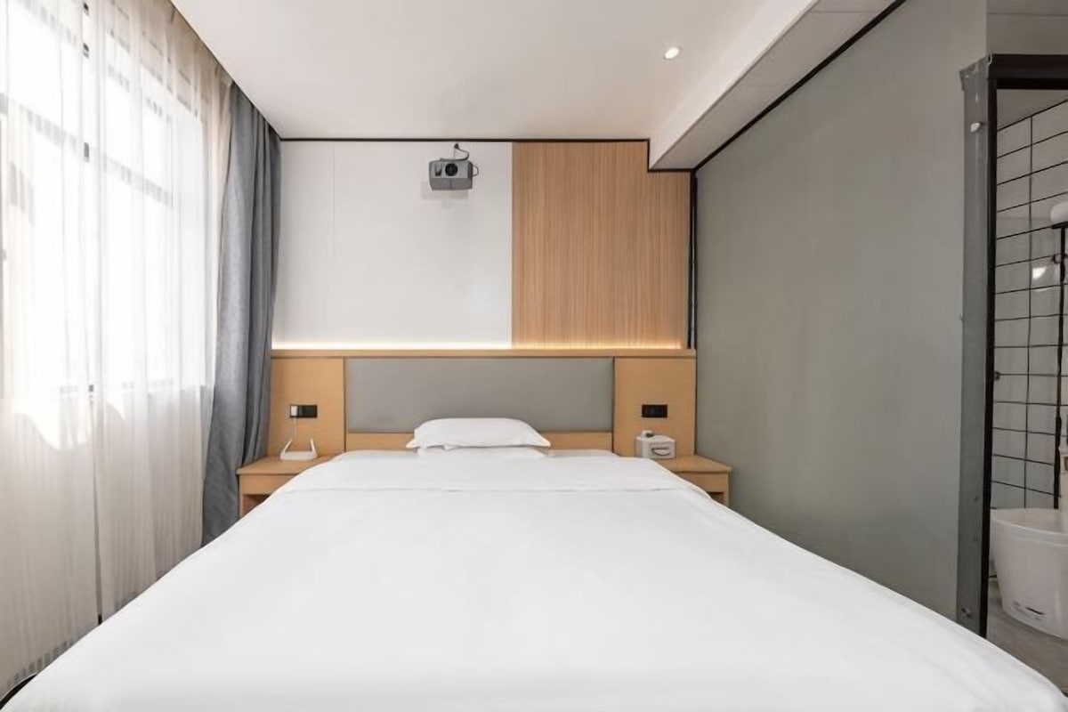 A modern hotel room with a large bed, bedside tables, a headboard with built-in lighting, and a window with curtains.