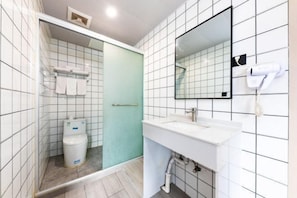 Un baño moderno con azulejos blancos, cabina de ducha de vidrio, lavamanos blanco con espejo y secador de pelo en la pared.