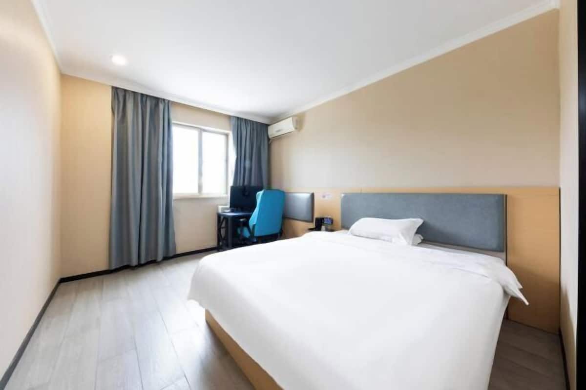 A hotel room with a large bed, a desk with a chair, a window with curtains, and an air conditioning unit on the wall.