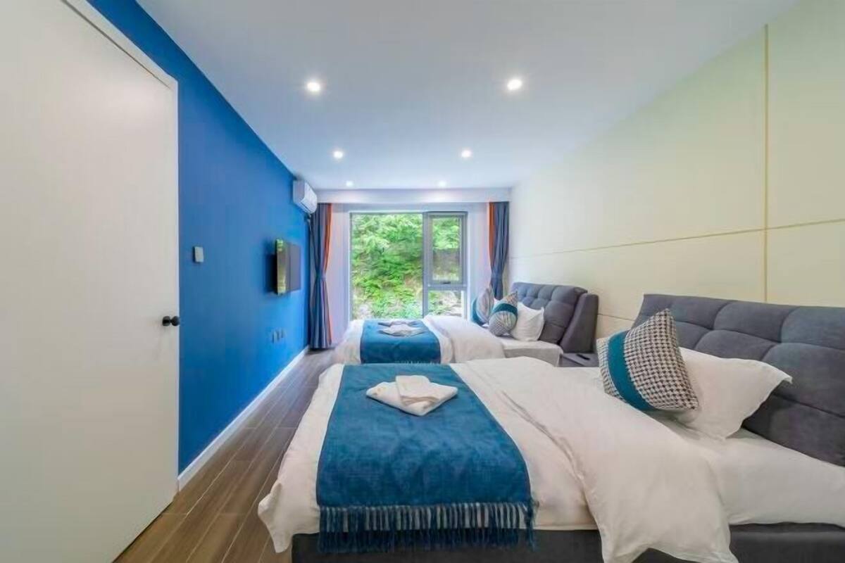 Quarto de hotel moderno com duas camas, uma janela grande com cortinas e vista para o verde lá fora.