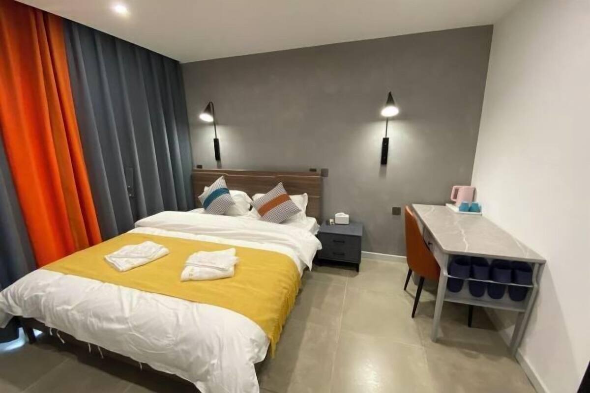 Quarto de hotel com cama, mesa de cabeceira, escrivaninha e prateleiras.