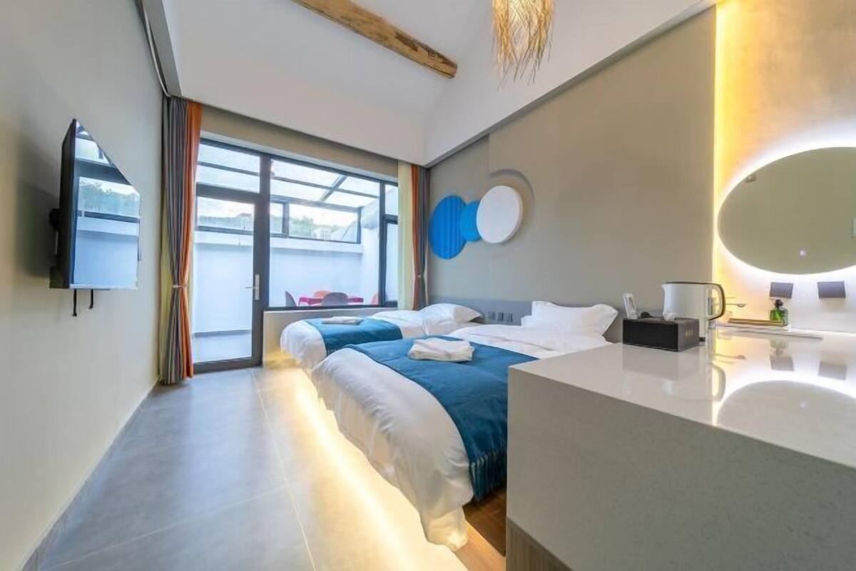 Quarto de hotel com duas camas, uma janela grande, televisão de tela plana, banheiro moderno com espelho e uma viga de madeira no teto.