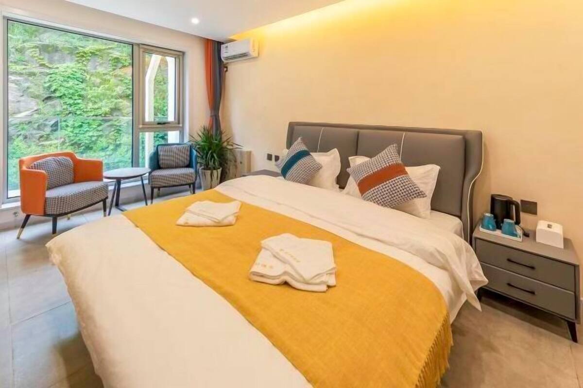 Quarto de hotel com uma cama grande, uma mesa de cabeceira, uma cadeira e vista para o verde pela janela.