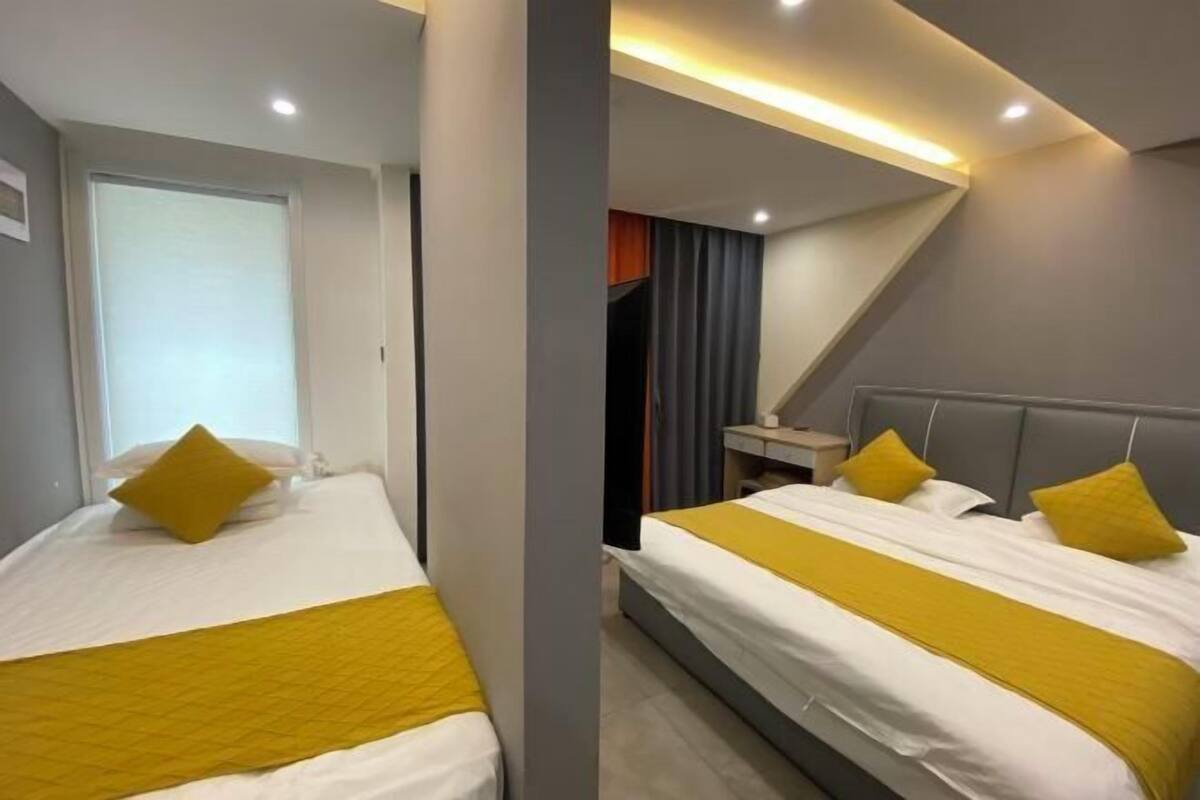 Quarto de hotel com duas camas, cada uma com colcha e almofadas amarelas, uma escrivaninha com cadeira e uma janela com cortinas.