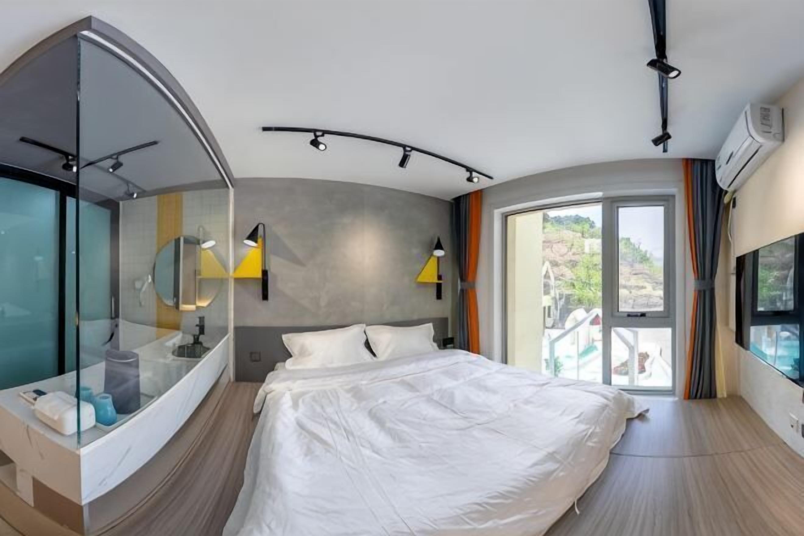 A modern hotel room with a curved glass wall, a large bed, a built-in vanity, and a view of a pool.