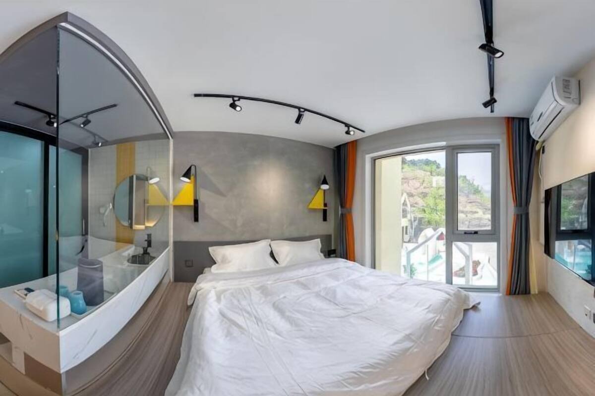 Quarto de hotel moderno com parede de vidro curva, uma cama grande, toucador embutido e vista para uma piscina.