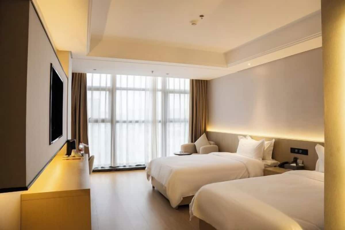 A hotel room with two beds, a desk, a flat-screen TV, and a large window with curtains.