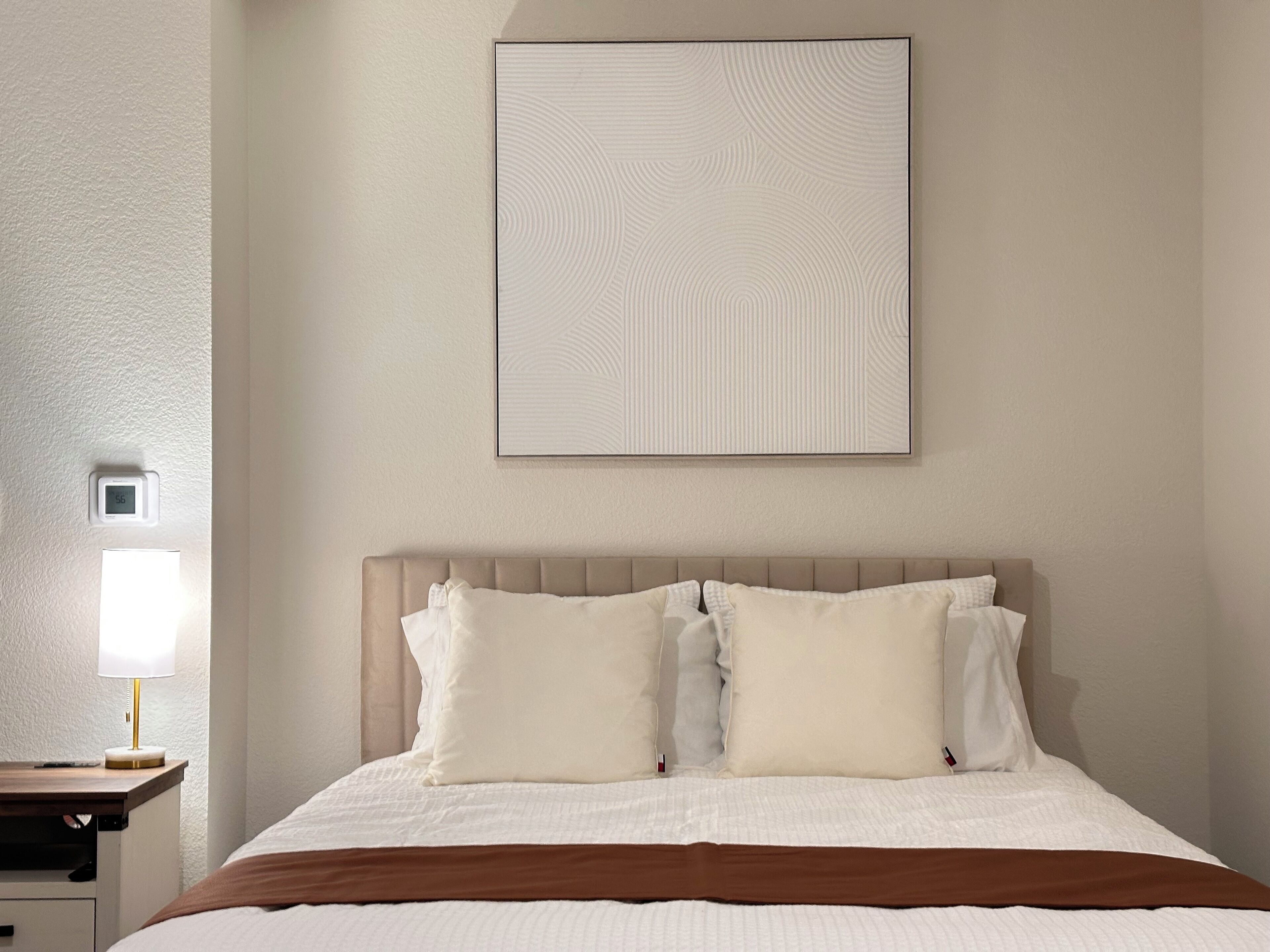 Una cama bien hecha con una colcha marrón, cabecera y dos almohadas. Se observa una mesita de noche con una lámpara y un pequeño reloj.