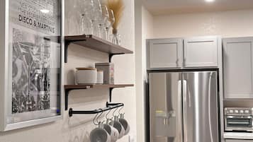 Een keuken met roestvrijstalen apparatuur, waaronder een koelkast en een oven. Er staan planken met wijnglazen en aan de muur een krantenartikel in een lijst.