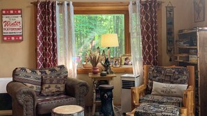 A cozy living room with a fireplace, a wooden coffee table, a sofa, and a chair. There is a window with curtains, a lamp, and a sign on the wall.