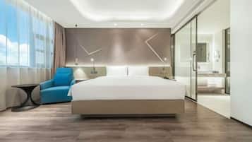 A modern hotel room with a large bed, a seating area, a glass partition, and a view of the sky.