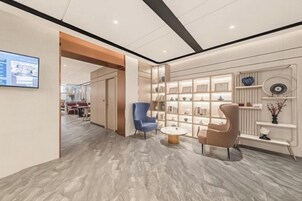 A modern hotel lobby with a marble floor, a blue armchair, a beige armchair, a small round table, and a wall with shelves displaying decorative items.