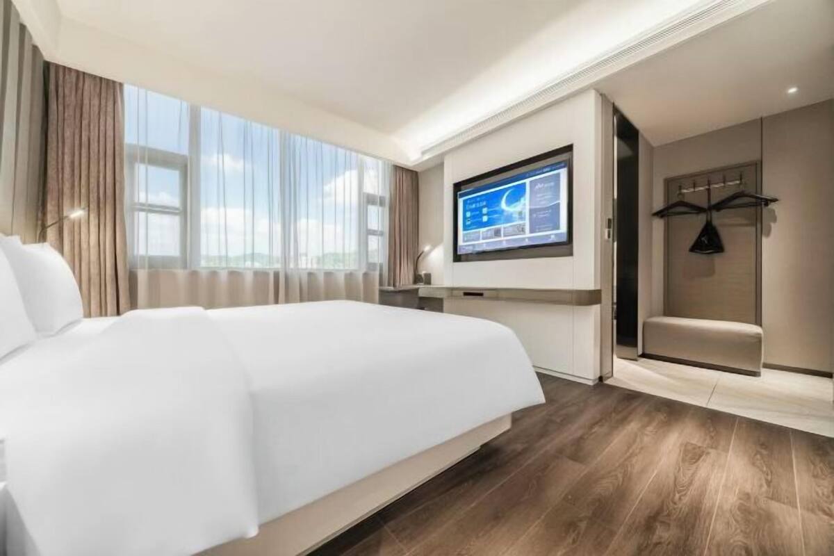 A modern hotel room with a large bed, a flat-screen TV mounted on the wall, a seating area with a bench, and a wooden floor.