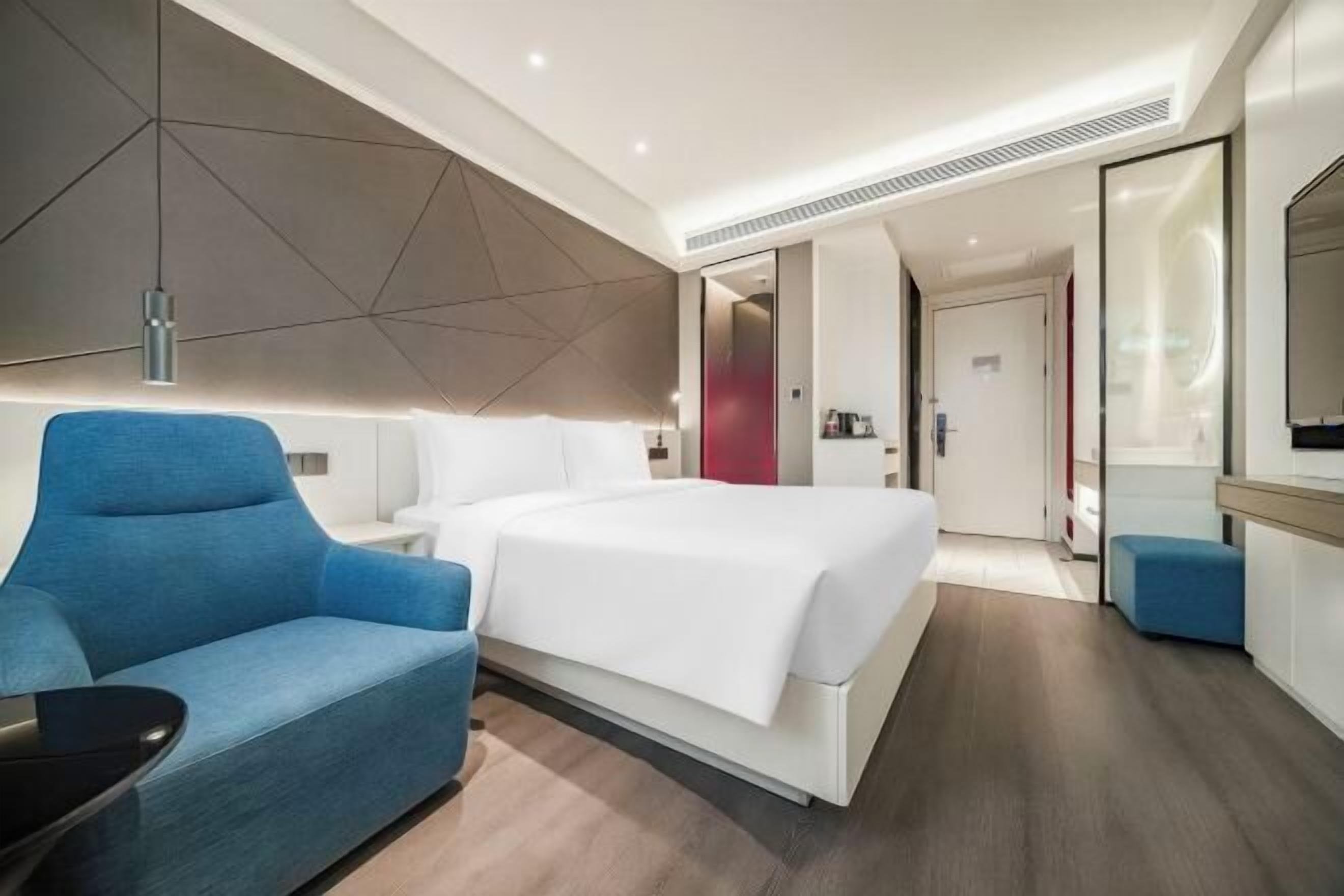 A modern hotel room with a large bed, a blue armchair, a bedside table, and a mirror.