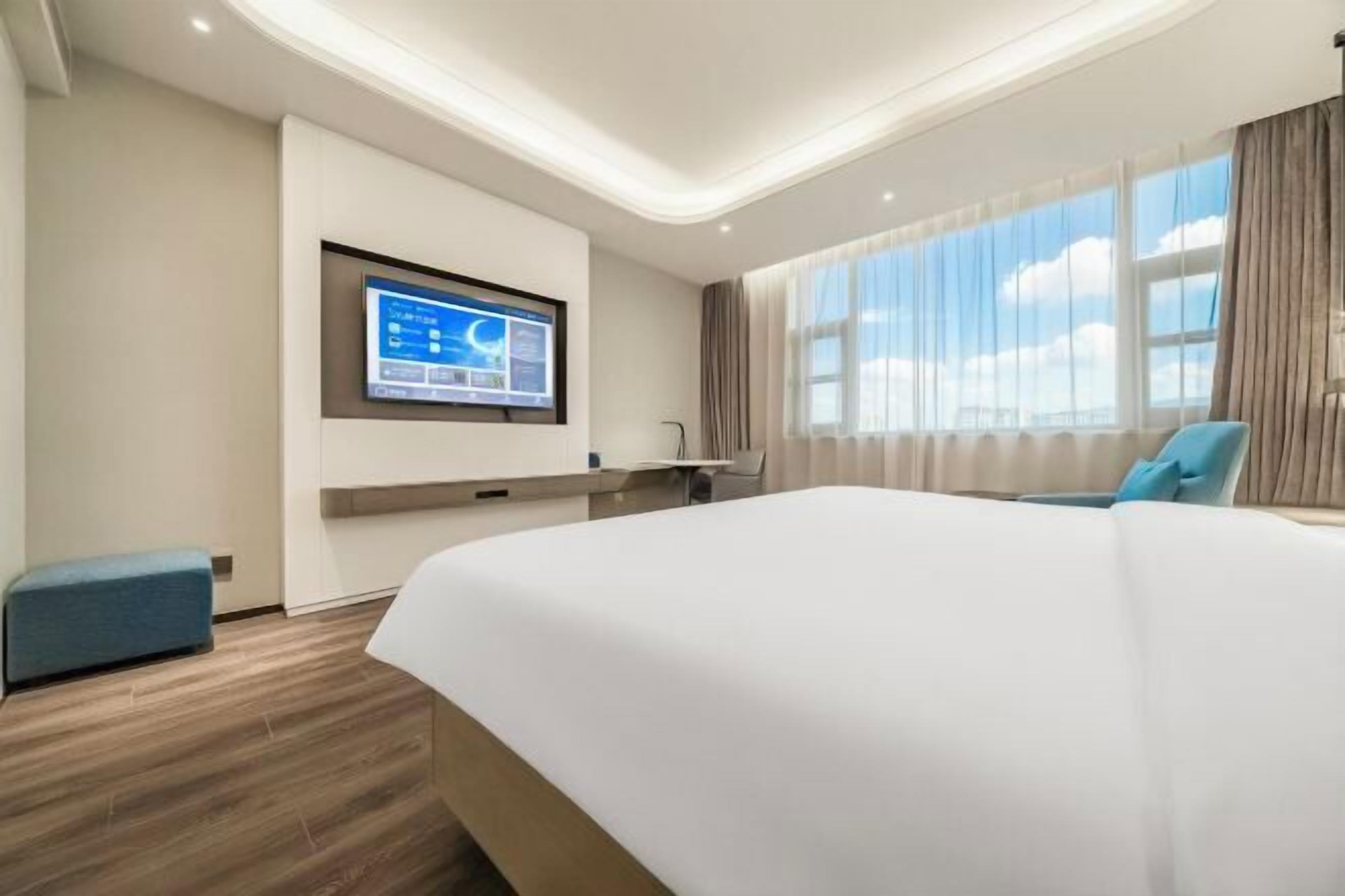 A hotel room with a large bed, a desk with a computer, a TV mounted on the wall, and a window with curtains.