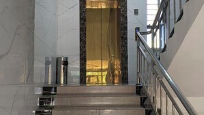 Un escalier en marbre avec une rampe métallique, menant à la porte d’un ascenseur en verre.