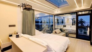 A modern hotel room with a large bed, a bedside table, a desk, a large window with a view, and a skylight.