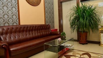 A brown leather sofa, a glass coffee table, and a potted plant in a room with patterned wallpaper and a tiled floor.