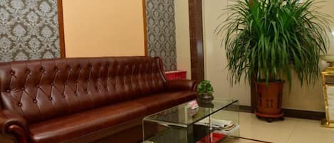 A brown leather sofa, a glass coffee table, and a potted plant in a room with patterned wallpaper and a tiled floor.