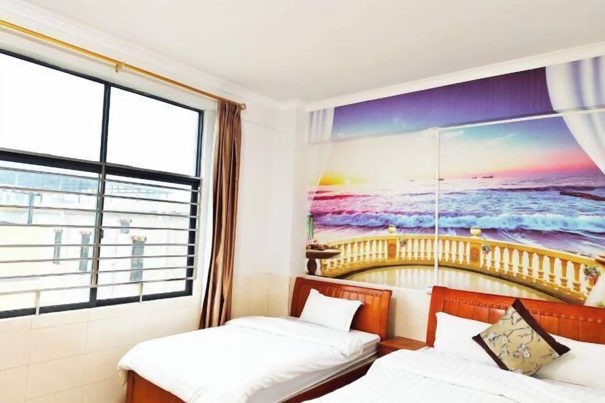 A hotel room with two beds, a large window with blinds, and a mural of a sunset over the ocean.