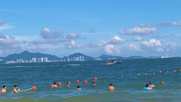 A coastal scene with people swimming and boats in the water, a clear sky, and distant buildings.