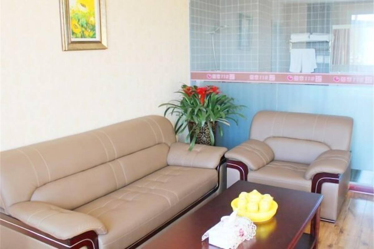 Una habitación con un sofá color beige, una mesa de centro de madera con una caja de pañuelos y un cuenco amarillo, y una planta en maceta con flores rojas.