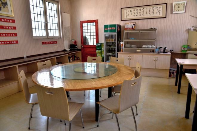 A room with a round glass table surrounded by chairs, a kitchen area with cabinets and a refrigerator, and a window with a view.