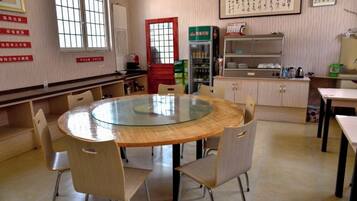 A room with a round glass table surrounded by chairs, a kitchen area with cabinets and a refrigerator, and a window with a view.