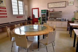 Una habitación con una mesa redonda de vidrio rodeada de sillas, una zona de cocina con armarios y un refrigerador, y una ventana con vistas.