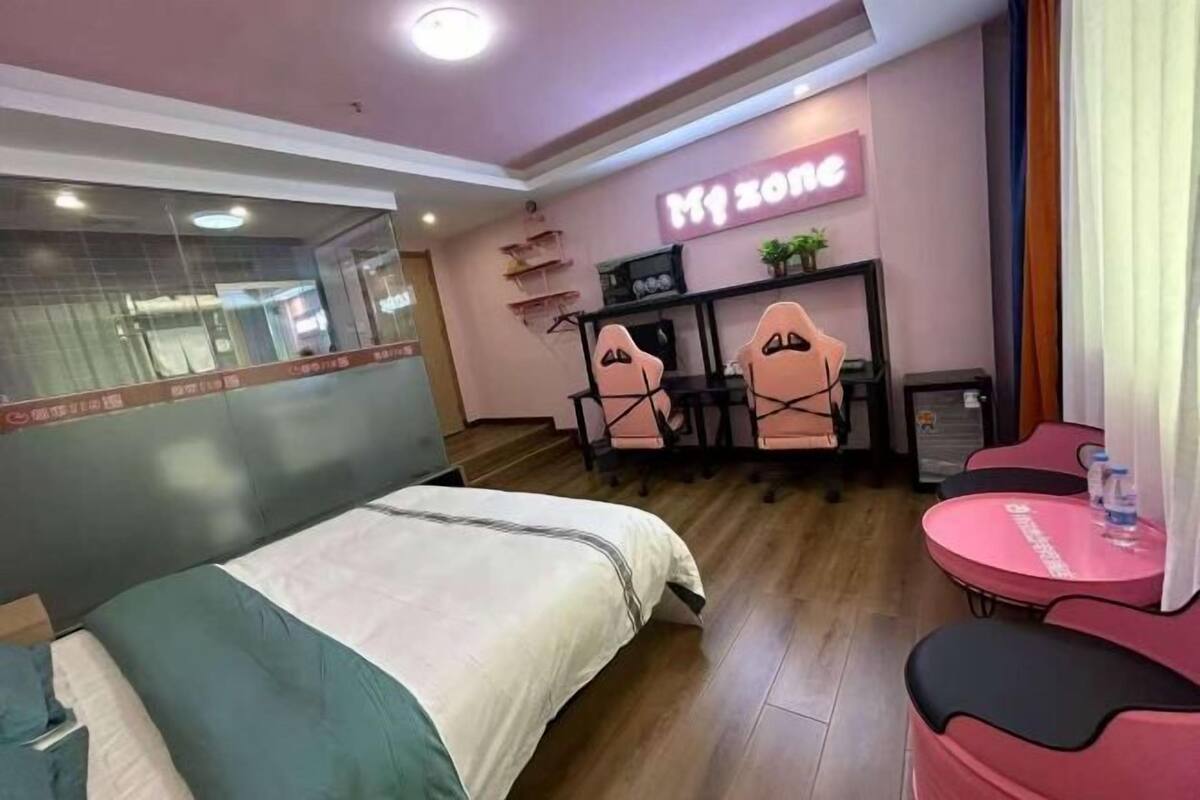 Habitación de hotel con cama, escritorio, dos sillas, una mesita con una botella de agua y un letrero de neón en la pared.