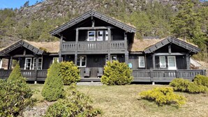 A two-story wooden cabin with a balcony, surrounded by well-maintained bushes and a mountain backdrop.