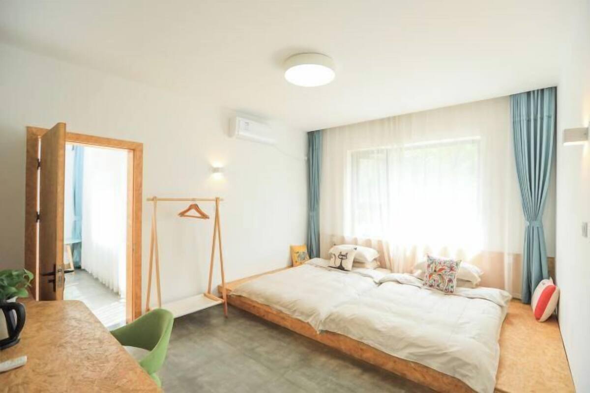 A bedroom with a bed, a wooden table, a chair, a hanging rack, and a window with curtains.
