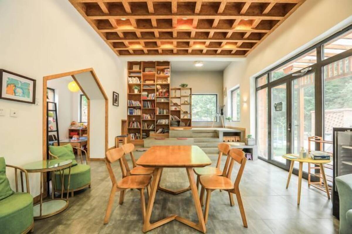 A modern dining area with a wooden table and chairs, surrounded by bookshelves and a view of the outdoors.
